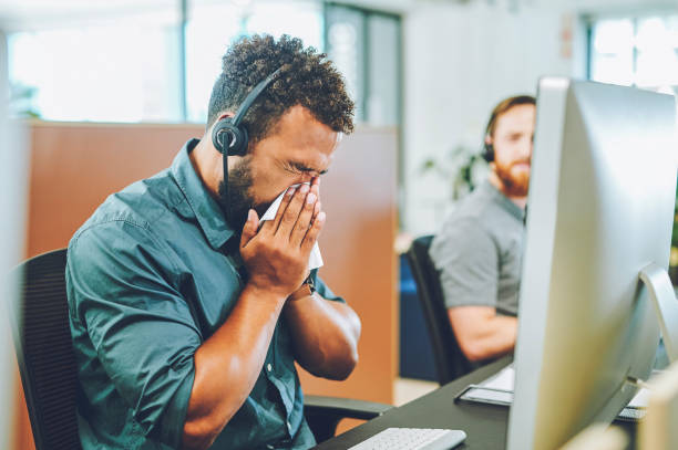 Sick, covid and unwell call center agent blowing his nose and spreading germ in an office space. An African customer service and support agent suffering from a cold or flu symptom at work stock photo