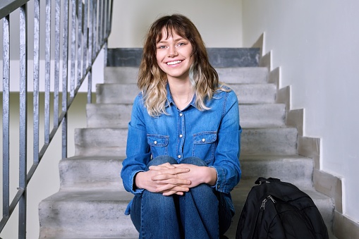 Portrait of smiling student girl with backpack on stairs inside building. Positive beautiful confident female with crossed arms looking at camera. Youth, education, lifestyle, young people concept