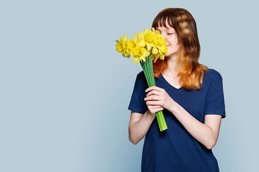 Young teenage red-haired female with bouquet of yellow flowers on light gray color background, teenage girl smelling flowers, copy space. Spring, beauty, holidays, youth concept