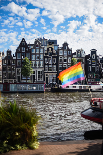 LGBT+ pride flag waving on a boat in a canal in the city of Amsterdam, the Netherlands. In the background several traditional Dutch buildings. Gay pride celebrations concept. Vertical image