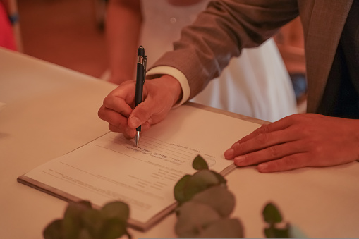The groom in a cream suit at the wedding reads the marriage contract, the bride stands next to her in a white dress, only the groom's hands with a handle are visible. On a white table lie roses with green petals. Front view. Selective focus.