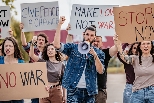 Group of people on strike on the street against war. Holding placards and posters. Young man talking into the megaphone.