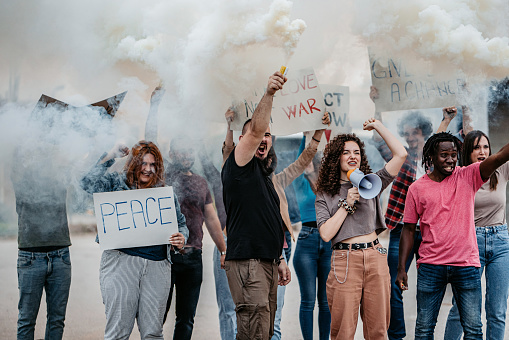 Group of people on strike on the street against war. Holding placards, posters and smoke bombs. Young woman talking into the megaphone.