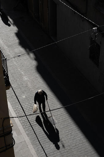 Top view of an old man with a stick walks on the street. Shadow in front of him. Man holding a shopper on a narrow street with residential apartments.