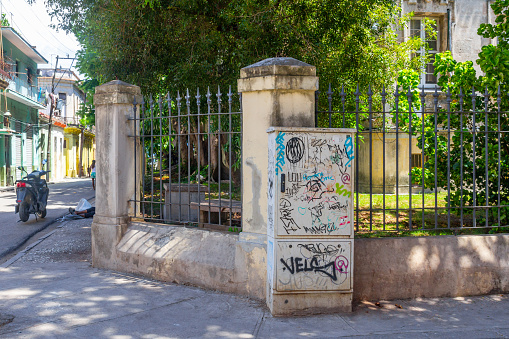 Havana, Cuba - July 28, 2022: A fenced house on a narrow street. A signpost painted with graffiti stands beside the fenced house.