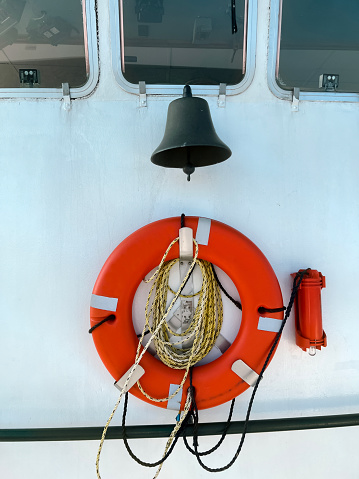 Closeup views of a bright orange life buoy hanging on the side of a passenger ferry boat. Nautical style ropes hang from the circle of life. A ship’s bell hangs above the life preserver. Attached to the wheelhouse of the ship.
