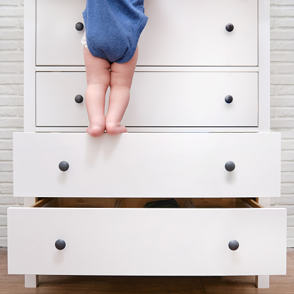 Toddler baby climbed up on the open chest of drawers. Child boy stood on a tall drawer of a white cabinet. Kid age one year