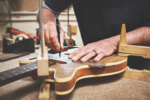 Closeup of man hands repairing, fixing and building a guitar in Luthiers service workshop. Professional male standing,  measuring and servicing music instrument on workbench in his garage.