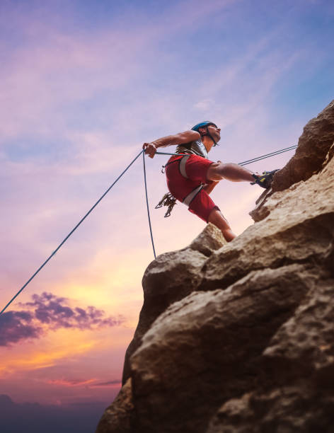 hombre escalador musculoso con casco protector haciendo rápel desde la pared de roca del acantilado usando el dispositivo belay de cuerda y el arnés de escalada en el fondo del cielo al atardecer. concepto de gasto de tiempo en deportes extremos activos. - rápel fotografías e imágenes de stock