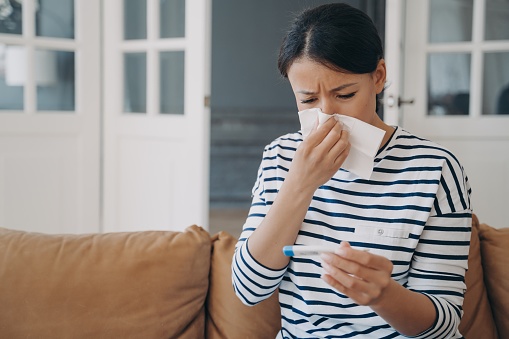 Sick unhealthy woman blowing nose checks temperature, suffering from influenza flu or cold sitting on sofa at home. Ill female looks at electronic thermometer, frowning, wiping runny nose with napkin.