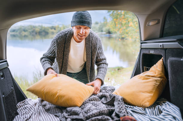 istock Midle-aged smiling man dressed in warm knitted clothes fixing pillows in opened Car trunk after a night on the beautiful autumnal mountain lake bank. Warm early autumn auto traveling concept image. 1414432228