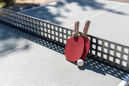 Two table tennis rackets leaning against a metal net and ping pong on a stone table outside on a sunny summer day.
