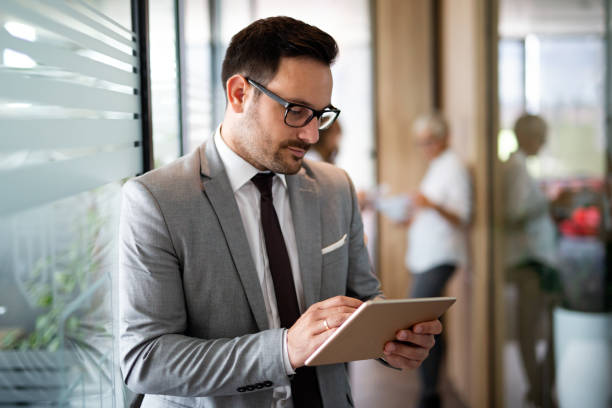 istock Attractive young businessman using a digital tablet while standing in front of windows in office 1414436683