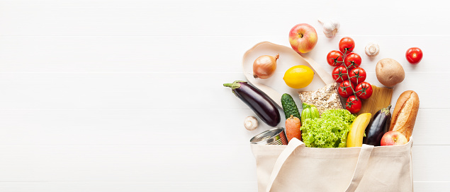 Cotton grocery tote bag with fresh vegetables, fruits, baguette and canned good on wooden background. Healthy food shopping, Eco-Friendly concept. Flat lay.