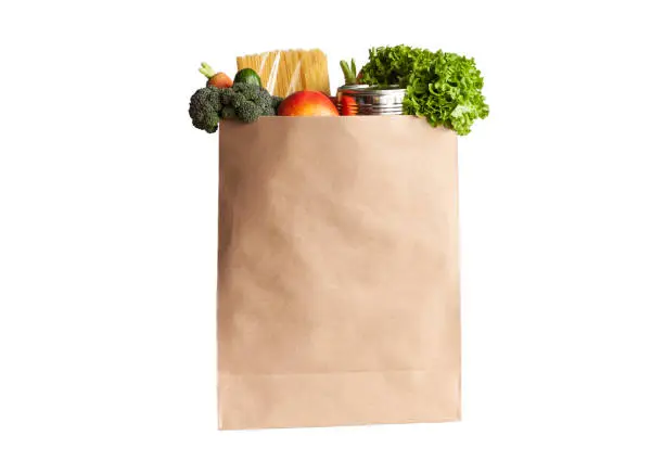 Paper shopping bag with various grocery items on white background, isolated. Bag of food with fresh vegetables, fruits, pasta and canned goods. Food delivery, shopping or donation concept.