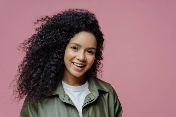 de cerca portrair de la joven modelo mujer sonriente tiene el pelo rizado, piel saludable, mira directamente a la cámara, expresa emociones positivas, disfruta de ser fotografiada, vestida con ropa de moda - frizzy 20s adult african descent fotografías e imágenes de stock