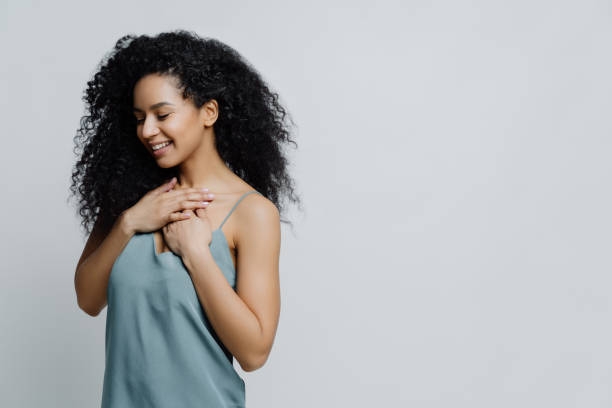 perfil de mujer étnica romántica de ensueño con corte de pelo afro, vestido con pijama, mantiene las manos en el pecho, recuerda momento muy agradable en la vida sonríe tiernamente con los ojos cerrados expresa sentimientos veraces - sólo adulto fotografías e imágenes de stock