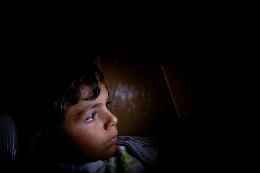 A 5.6-year-old boy shines the light of the table on his side-plan face in the dark environment. He looks closely at the screen of the tablet with all his attention.