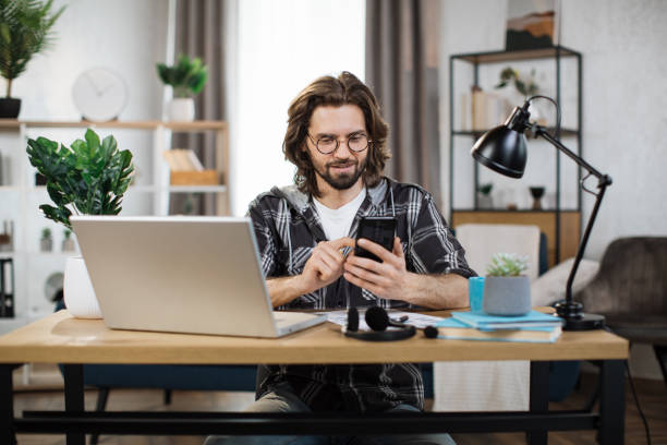istock Man in casual outfit typing on mobile phone while sitting at workplace with laptop. 1414451458