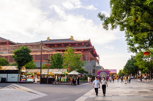 Xi'an, China - July 29, 2022: People walking along the walkway in a busy tourist area. There are some kiosks and a large traditional Chinese building in the scene.