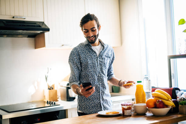 istock Smiling man having breakfast in modern kitchen and using smart phone for online messaging or e-banking 1414456280