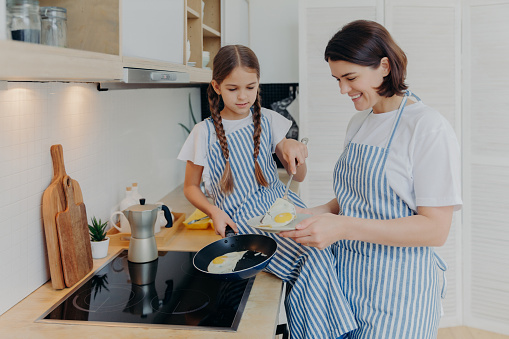 Photo of cheerful busy mum and her daughter pose near stove, serve breakfast for family, fry eggs on frying pan, wear aprons, pose in domestic kitchen. Mother teaches kid how prepare meal easily
