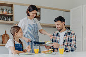 istock Busy housewife holds frying pan, gives prepared meal to husband and daughter, have delicious breakfast together, sit at kitchen table with glass of juice and sandwich. Happy family eat at home 1414458102