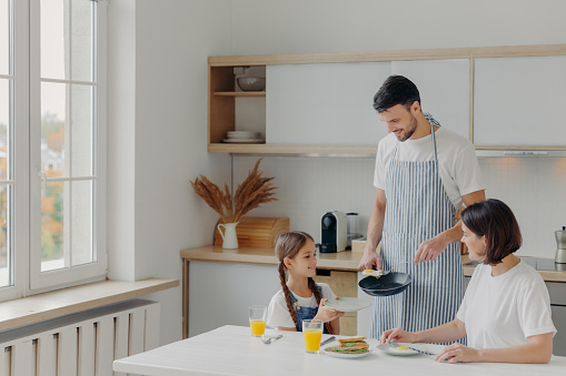 Father prepared fried eggs for family, little child holds plate and waits for breakfast. Family pose at kitchen near table, enjoy tasty meal, have glad expressions. People, eating, domestic atmosphere