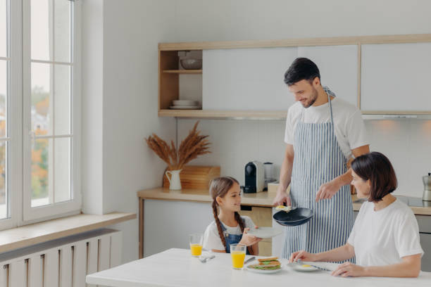 pai preparou ovos fritos para a família, criança segura prato e espera pelo café da manhã. família posa na cozinha perto da mesa, desfruta de refeição saborosa, tem expressões alegres. pessoas, comida, atmosfera doméstica - stereotypical housewife little girls family domestic kitchen - fotografias e filmes do acervo