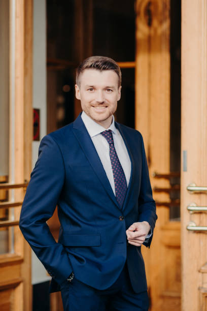istock Attractive male executive manager in formal outft, keeps hand in pocket, has positive expression, stands near office, rejoices good achievement, confident in success. Male lawyer stands near doors 1414459865