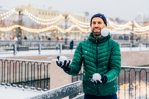 Playful smiling male juggles with snowballs throws them in air, has happy expression, stands against beautiful winter backgrounds. Happy overjoyed male model has fun outdoors, enjoys snowy views