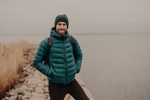 Horizontal shot of attractive unshaven man has cheerful expression, wears hat and jacket, keeps hands in pocket, carries rucksack, has walk during foggy autumn weather, poses near lake outdoor