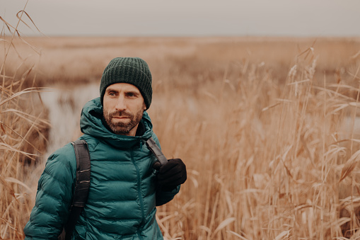 Horizontal shot of contemplative good looking man has stubble, wears hat, jacket and gloves, stands near wheat field background with free space on right side. People, leisure and walk concept
