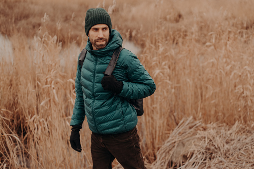 People and journey concept. Shot of handsome man dressed in warm jacket and headgear, walks outside, looks attentively aside, poses against field background with copy space on left side for text