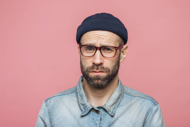 istock Grumpy bearded man with offended expression, being dissatisfied with something, frowns face, wears spectacles, hat and shirt, isolated over pink background. Displeasure and negativity concept 1414464598