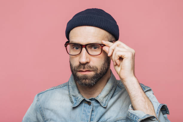 istock Handsome bearded man with serious expression wears spectacles and glasses, dressed in denim fashionable shirt, isolated over pink background. People, facial expressions and emotions concept. 1414464600