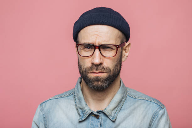 istock Unhappy discontent unshaven male looks with grumpy expression into camera, wears glasses black hat and stylish denim shirt, isolated over pink background. Displeased middle aged man poses indoor 1414464605
