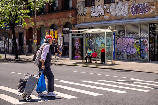 Lower Manhattan, New York, NY, USA - July 4 2022: Man with a shopping chart crossing the Bowery in a zebra crossing in front of a bus stop on a sunny summer day