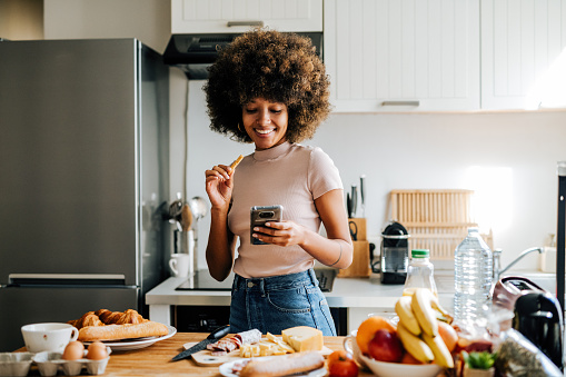 Mujer afroamericana de cabello rizado en casa en la cocina, preparando el desayuno y usando un teléfono inteligente photo