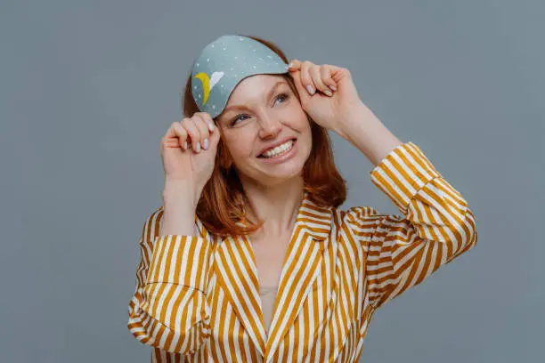 Dreamy redhaired European woman looks gladfully aside, smiles broadly, wears casual domestic clothes and sleepmask, isolated over grey background, imagines something pleasant. Good morning concept