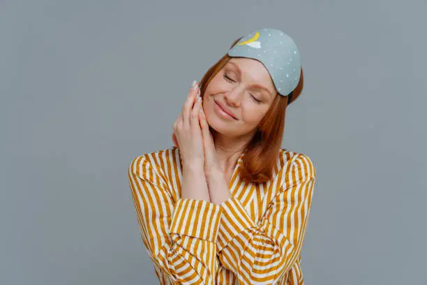 Adorable freckled red haired woman keeps eyes closed, hands together near freckled face, wears sleepmask and casual pajama, isolated over grey background, has pleasant dreams during afternoon nap.