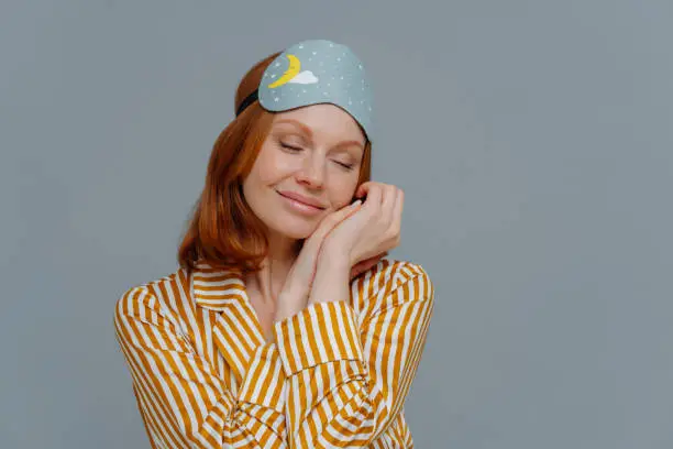 Pretty foxy freckled woman leans at palms, keeps eyes closed, wears sleepmask and stiped pajamas, dreams about something pleasant, stands indoor over grey wall, with copy space for promotion