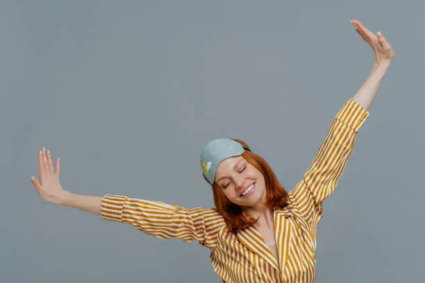 Perfect rest and sleeping concept. Cheerful young ginger European woman spreads arms and stretches after awakening, had sweet dreams, wears striped pajama and sleepmask, isolated on grey wall