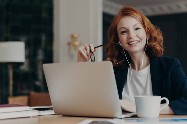 istock Young happy female freelancer with ginger hair thinks about new ideas for work, works distantly on laptop computer, prepares for video conference, uses earphones, poses at workplace with hot drink 1414481342
