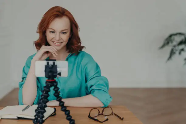 Happy young ginger woman shoots video in front of smartphone videocamera on tripod, gives recommendations how to start own business, has personal vlog, speaks on video call about online training