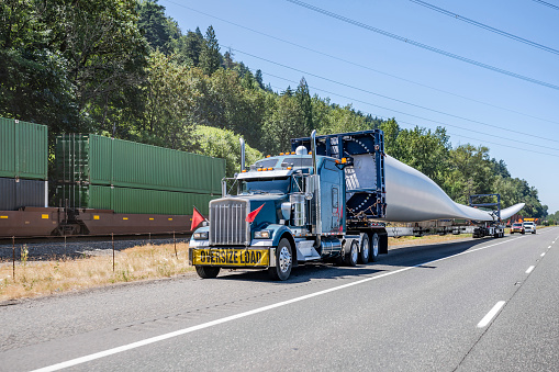 Powerful Big rig semi truck tractor with oversize load sign transporting windmill electric generator blade with special additional trolley standing on the highway shoulder with escort vehicles