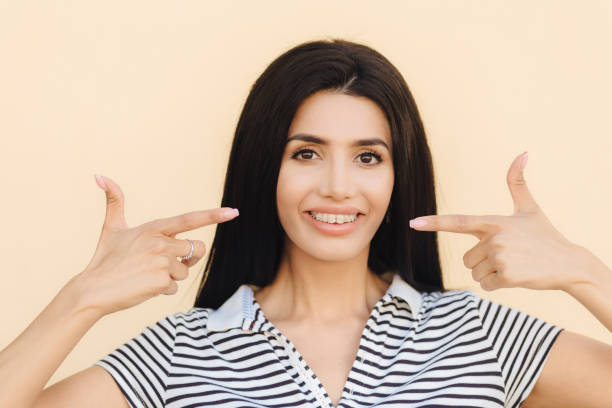 istock Portrait of pleasant looking brunette female has appealing look, indicates with both fore fingers at mouth shows gentle smile, teeth with braces, dressed in casual clothes, isolated on light wall 1414482096