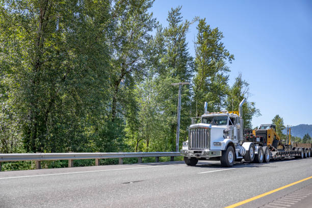 istock Powerful white classic day cab big rig semi truck transporting oversize load on the step down semi trailer driving on the road in sunny day 1414483190