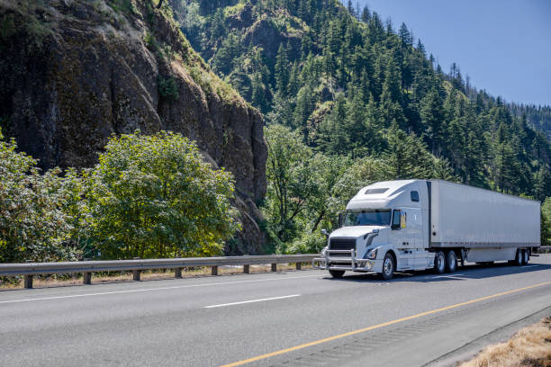 istock Powerful white big rig semi truck with grille guard transporting cargo in dry van semi trailer driving on the highway road with mountain and trees on the side 1414484582