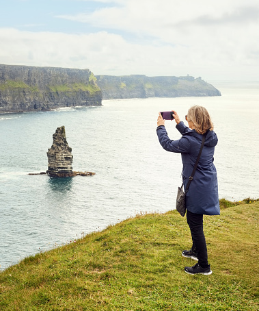 Rear view of a female tourist taking pictures of coastal cliffs with her mobile phone